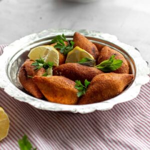 A close-up of golden, crispy kibbeh served with lemon wedges and fresh herbs on a silver platter.