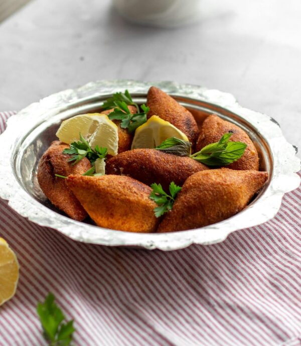A close-up of golden, crispy kibbeh served with lemon wedges and fresh herbs on a silver platter.